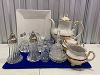Full view of porcelain tea set with sugar bowl, creamer, three sets of glass salt and pepper shakers, square plate, glass butter dish, trinket container, and bowl all laid on blue cloth.
