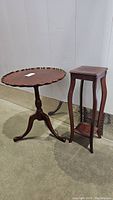 Photo displaying the vintage wooden pedestal table with scalloped edge and tripod legs next to the tall plant stand with curved legs and a lower shelf, both dark wood.