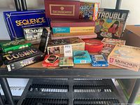 Photo showing a variety of board and card games stacked on a surface including Sequence, Rummikub, Yahtzee, Trouble, Cosmic, Munchkin, Uno, and others.