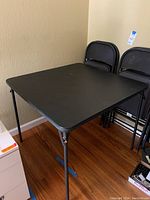 Black folding card table set against a beige wall with four matching folding chairs stacked behind it. The table has a square shape and metal legs.