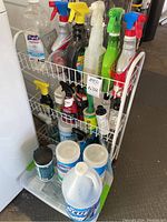 Overall view of white wire rack filled with assorted cleaning supplies including sprays, wipes, bleach, sanitizer.