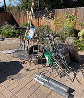 Wide view of lot showing metal plant stands, rakes, mulch bags, buckets, wire mesh rolls, irrigation tubing, and outdoor rugs on a brick patio.