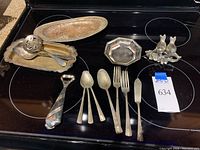 Photo showing small oval platters, serving spoons, plated cutlery including forks, spoons, and a knife, a distinctive tie-shaped bottle opener, and bird salt and pepper shakers arranged on a black surface.