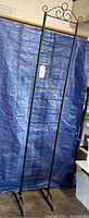 Full view of tall black metal quilt rack with blue tarp backdrop showing entire rack height and feet.