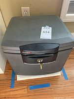 Closed dark gray Sentry Safe with keys inserted in lock, sitting on floor.