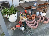Overview of outdoor garden supplies on concrete patio including pots, edging stones, extension cords, bird figurines, trivets, stool, side table, and various garden tools and accessories.