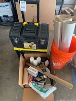 Closed Stanley toolbox on wheels, two orange Home Depot buckets containing rolled sheets, and cardboard box with painting supplies