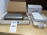 Photo showing ultrasonic cleaner in metallic tray with power cord beside a white rectangular PhoneSoap UV sanitizer in their boxes.
