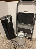 Black Holmes air purifier tower next to the step ladder and humidifier on carpeted floor with white backdrop.