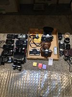 Overview of film cameras, lenses, film canisters, meter, flash and binoculars arranged on floor