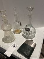 Three glass decanters, two with metal tags, and one metal flask on white surface.