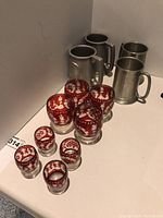 Photo showing six red decorated glasses with ornate floral cut designs and four pewter beer tankards with handles on a flat surface.