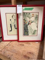 Two framed artworks side by side on wooden surface, red frames, one depicting a woman in gloves, the other a mounted horseman.