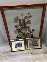 Three framed pictures arranged on the floor: one large needlepoint picture with wine and grape theme, two smaller gold framed silk pictures showing scenic views.