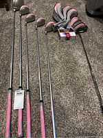 Five Lopez golf drivers numbered 1,4,5,6,7 with pink grips and head covers, two sleeves of golf balls on carpeted floor