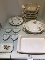 Photo showing three stacked CorningWare casserole dishes with lids, porcelain serving bowl with lid, four heart-shaped cheese strainers, decorative serving plate with scalloped edges, and rectangular baking dish.