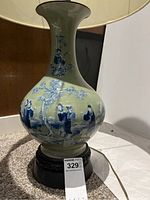 Close-up view of ceramic lamp base with hand painted Asian-style blue and white artwork showing people and trees. Mounted on a dark round wooden base.