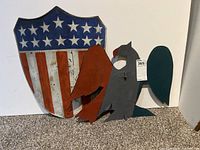 Front view of the painted metal shield with American flag design alongside two bird-shaped metal props, one red and one black with green wings.
