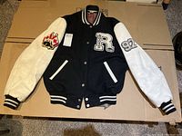 Full front view of black and white Roots varsity jacket laid flat showing front patches, pockets, and snap button closure.