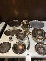 Full view of the collection of various silver-tone serving ware on a table, showing trays, covered dish, ladle, small dishes, and shaker, all with tarnishing and ornate detailing.