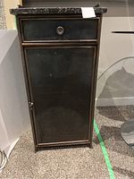 Front view of small metal cabinet showing drawer and door with metal knobs.