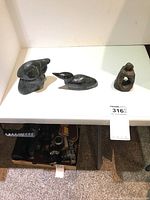 Overview photo showing all three statues on a white surface. Two are carved stone birds and one is a small metal figurine.