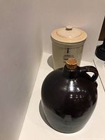 Photo showing both the bean pot with lid and the dark brown stoneware jug with cork stopper placed on a white surface.