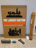 Mounted railroad memorabilia auction poster with steam locomotive graphic and orange and white text, two large rail spikes, two small metal replicas, and a rolled additional poster wrapped in brown paper, all on carpeted floor.