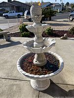 Overview front view of the full 3-tier outdoor fountain with multi-colored river rocks in basin, placed on concrete patio with plants and bench in background.