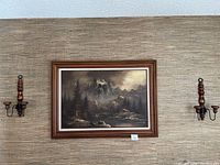 Framed oil painting depicting a mountain landscape flanked by two wooden wall-mounted candle holders.