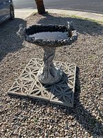 Full view of the birdbath placed on gravel with decorative concrete tiles underneath. The basin contains some water. It shows small bird sculptures on the rim and a textured edge.