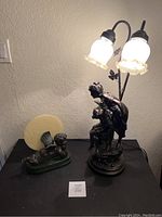 Two bronze-style lighting statues on a black surface; a cherub lamp on left with frosted round glass panel and a lamp sculpture of woman and child holding a basket with two lit glass lamps on right.