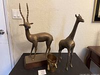 Photo showing three bronze/brass animal statues: large gazelle with long horns mounted on a base, a giraffe statue, and a small deer head figurine.