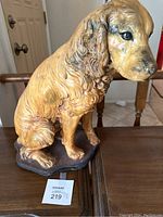 Front and side view of painted dog statue placed on wooden table with auction tag 219 visible