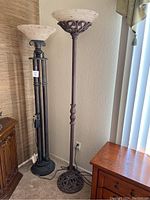 Two ornate metal floor lamps standing side by side with detailed bases and upward-facing speckled shades beside wood furniture and window blinds.