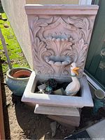 Full view of beige floral wall fountain with electrical cord, fountain basin holding duck figurine and miscellaneous small items, blue ceramic pot with terra cotta pot inside on ground next to fountain.