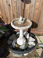 Side view of the concrete bird bath featuring a round basin and frog figurine on top, placed in plastic tub with rocks.