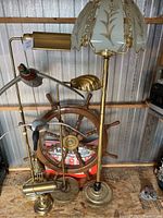Photo showing three floor lamps, including a tall lamp with etched glass floral shade, brass desk lamp, and nautical ship wheel in background.
