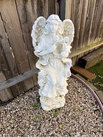 Full view of white angel statue holding a lyre, set against wooden fence and gravel ground.