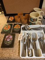 Overview photo showing silverware in tray, ceramic mugs, vintage-style glasses, and floral ceramic coasters and plaques arranged on countertop.