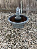 Photo of small gray stone-like outdoor fountain on gravel surface full of water with central column and floral decoration on base.