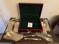 Gold tone silverware pieces including forks, spoons, knives, mostly still wrapped individually, spread around and inside the open wooden box with green velvet interior.