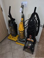 Photo of three upright vacuum cleaners: Dyson yellow, Bissell black, Eureka gray. Showing general condition and inclusion of hoses and power cords.
