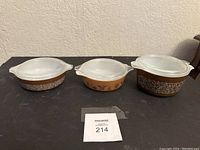 Three vintage brown Pyrex casserole dishes with their white lids, each with different patterns on their sides.