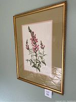 Framed botanical print wall art showing a pink snapdragon flower illustration with green foliage, gold frame, and multi-layered matte.