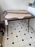 Full view of the vintage kitchen table showing the laminate woodgrain top with brown border and tan abstract shapes, starburst decorations near edges and metal legs.