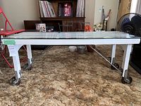 Wide angle side view of the luggage rack showing the full length with glass top, white painted wooden slats and caster wheels.