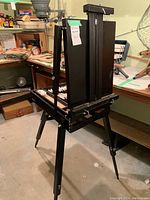 Black wooden portable art easel set up in basement/workshop area, showing easel structure, adjustable legs, and storage box beneath with clasp closures.