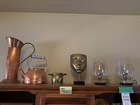 All items displayed on a wooden shelf: tall copper jug, copper kettle, small brass pot, brass face mask sculpture, and pair of brass candleholders with glass covers.
