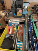 Wide view of drill bit and tap sets on wooden surface, large Bosch SDS bits in tube, assorted accessories and packaging.